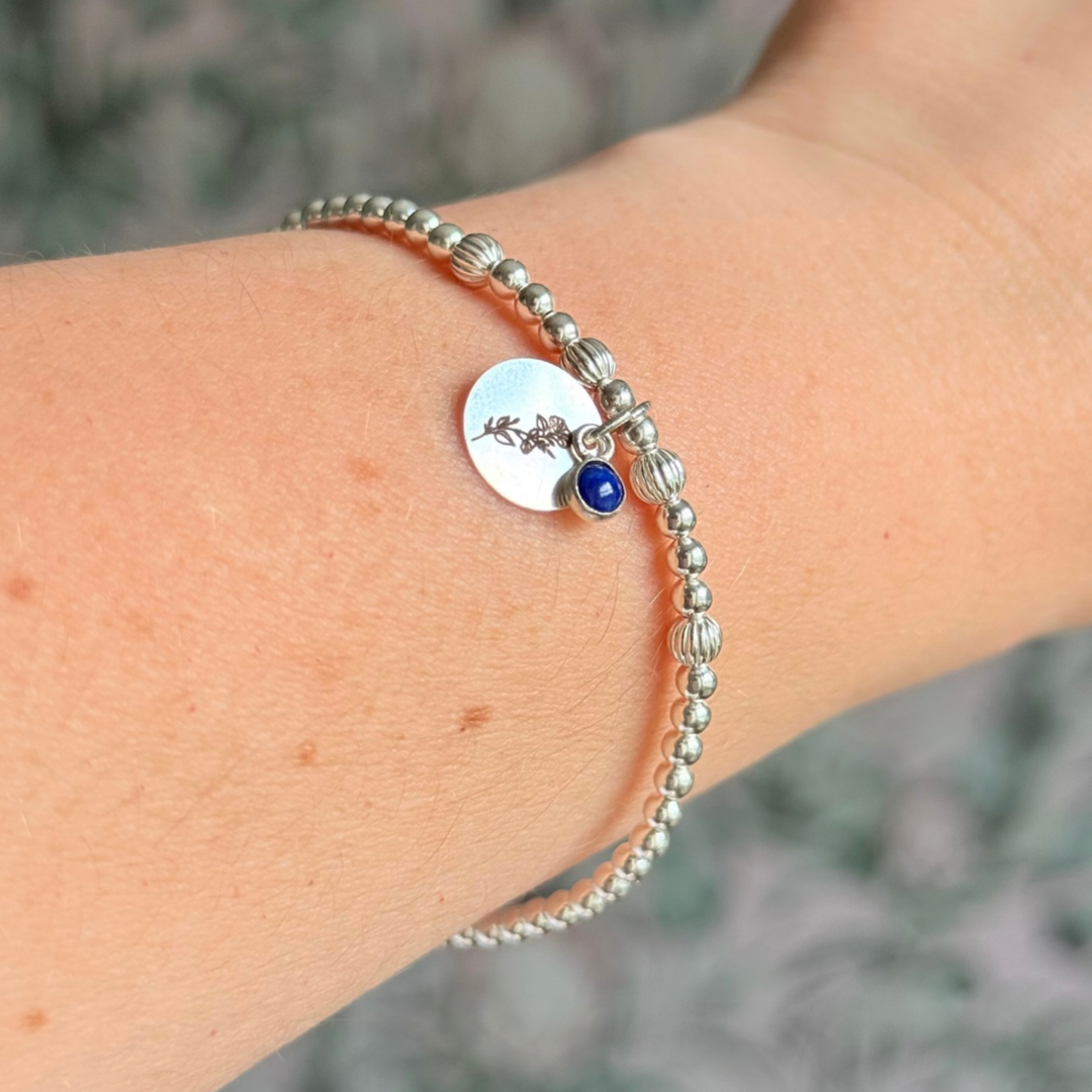 This sterling silver bracelet features a domed 4mm lapis lazuli cabochon charm, the birth stone for September, in a sterling silver setting. It features an 11mm sterling silver disc charm, which is engraved with the September birth flower, Morning Glory. This charm can be personalised on one side if required or feature the same flower graphic on both sides. The main band of the bracelet is made up of 3 mm sterling silver round ball beads with some corrugated accent beads.
