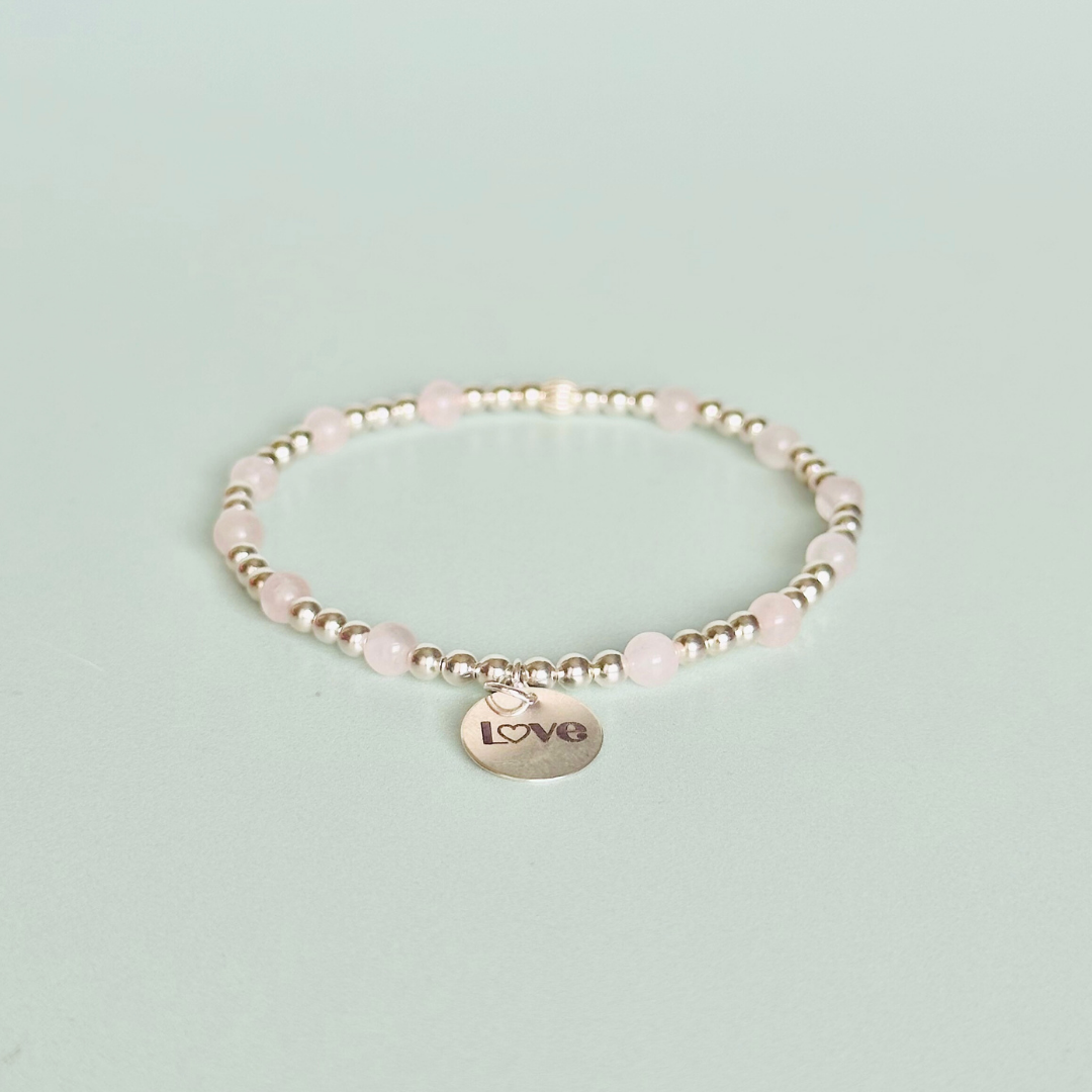 Sterling silver elasticated bracelet featuring 4mm round rose quartz beads, separated by 3mm sterling silver round ball beads. Featuring an 11mm sterling silver round disc charm engraved with the words love. One side of the charm can be personalised if required.
