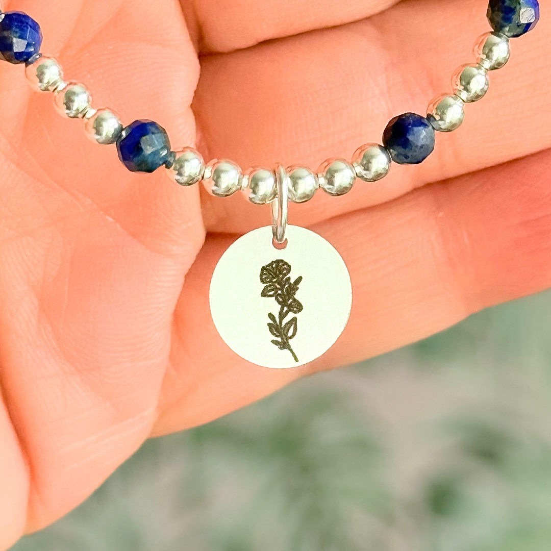 Sterling silver elasticated bracelet featuring 4mm faceted round lapis lazuli beads, separated by 3mm sterling silver round ball beads. Featuring an 11mm sterling silver round disc charm engraved with the September birth flower Morning Glory. One side of the charm can be personalised if required.