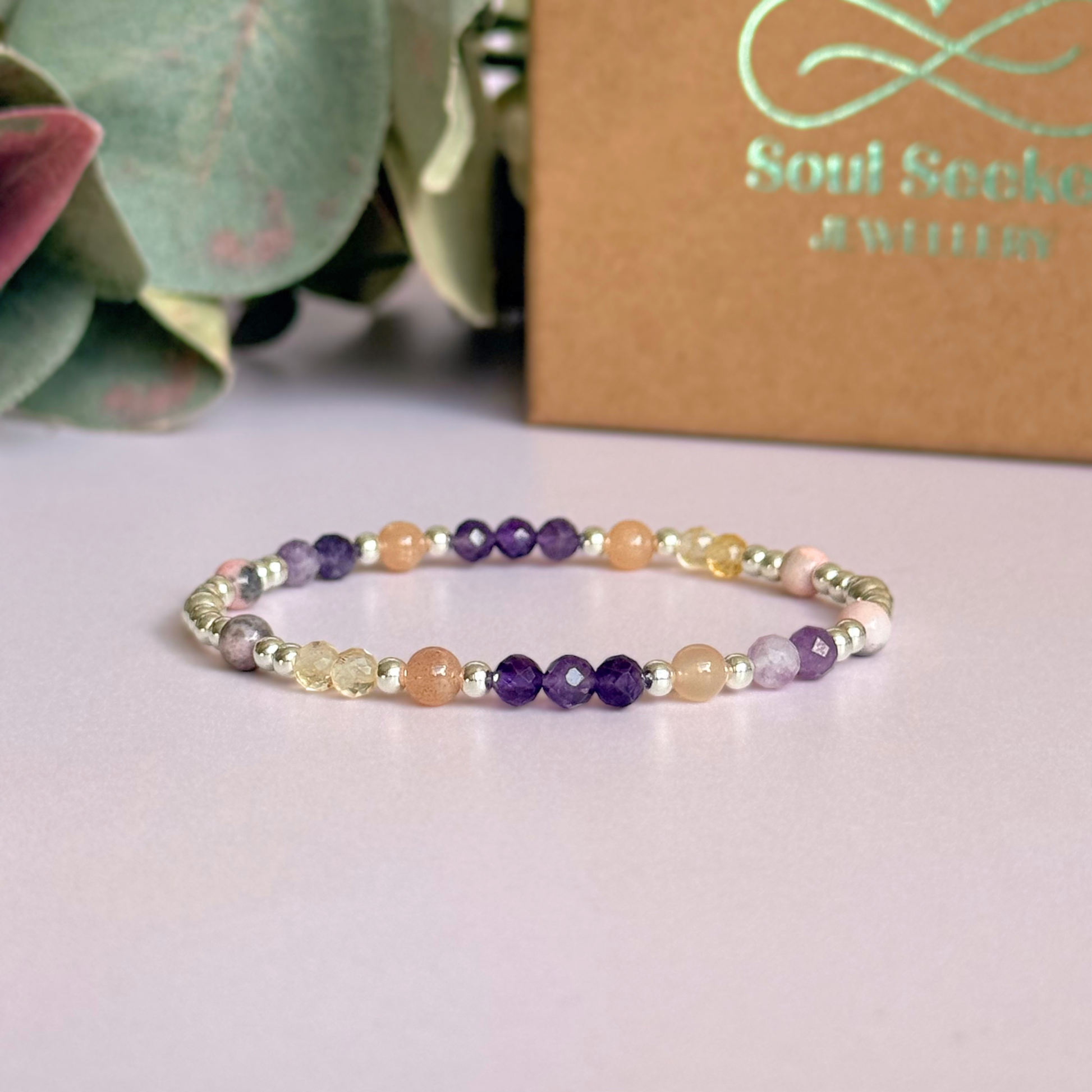 Dainty sterling silver crystal bracelet featuring five crystals, in 4mm faceted and smooth round style, each believed to help with menopause symptoms. Featuring amethyst, citrine, lepidolite, peach moonstone and pink zebra jasper. Pictured against a pink background with the box in the background.