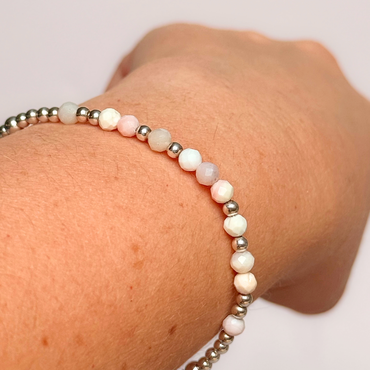 Dainty, minimalist sterling silver and pink opal October birthstone stacking bracelet. Made with 4mm faceted pink opal beads and 3mm sterling silver round beads. Elasticated for comfort. Pictured being worn, close up