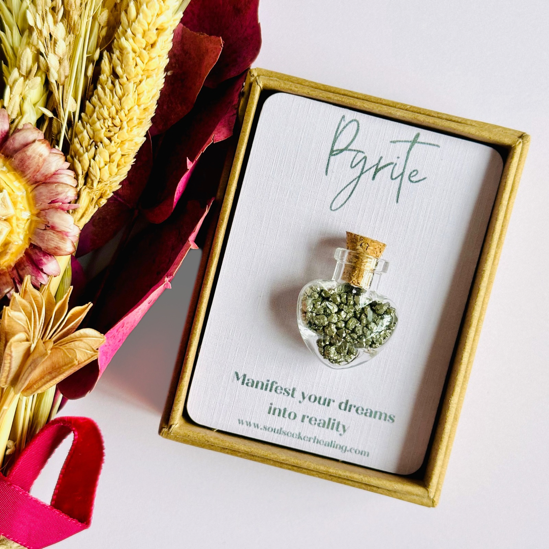 A pretty heart shaped mini glass bottle filled with powerful, ethically mined pyrite crystal chips, presented on an eco-friendly textured card with the words 'Manifest your dreams into reality.’