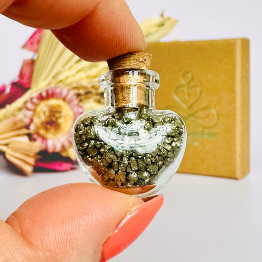 A pretty heart shaped mini glass bottle filled with powerful, ethically mined pyrite crystal chips, presented on an eco-friendly textured card with the words 'Manifest your dreams into reality.’