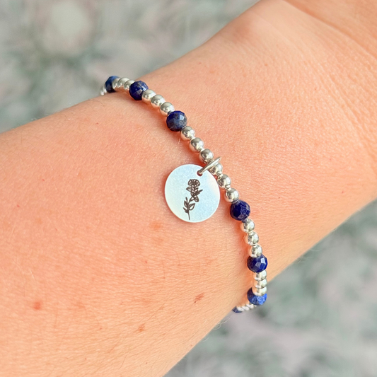 Sterling silver elasticated bracelet featuring 4mm faceted round lapis lazuli beads, separated by 3mm sterling silver round ball beads. Featuring an 11mm sterling silver round disc charm engraved with the September birth flower Morning Glory. One side of the charm can be personalised if required.