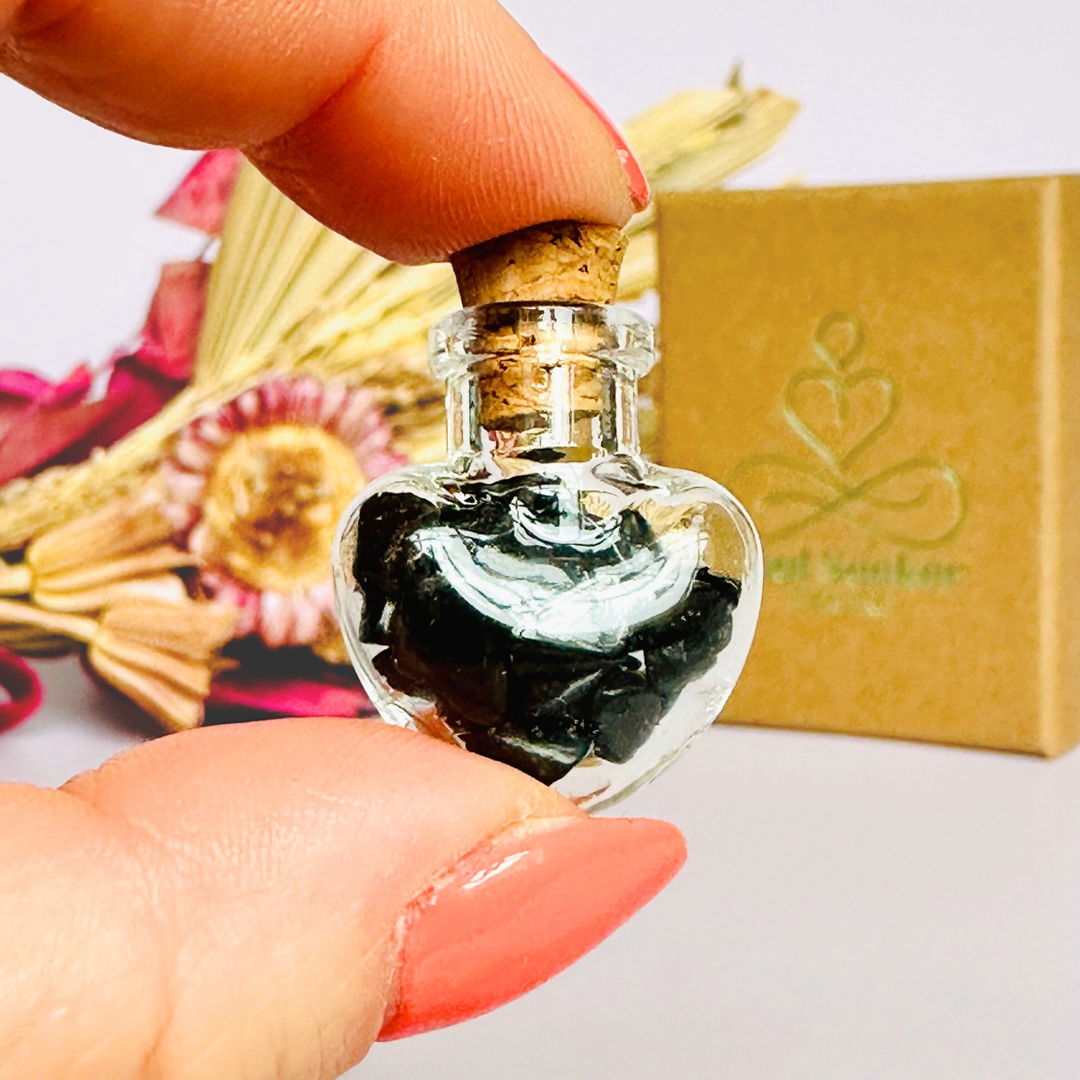 A pretty heart shaped mini glass bottle filled with powerful, ethically mined black tourmaline crystal chips, presented on an eco-friendly textured card with the words 'Protection against negative energy.’