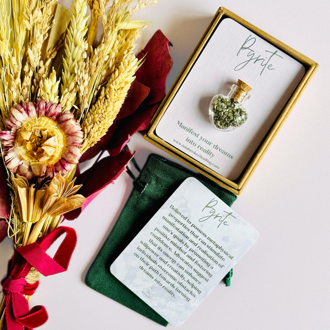 A pretty heart shaped mini glass bottle filled with powerful, ethically mined pyrite crystal chips, presented on an eco-friendly textured card with the words 'Manifest your dreams into reality.’