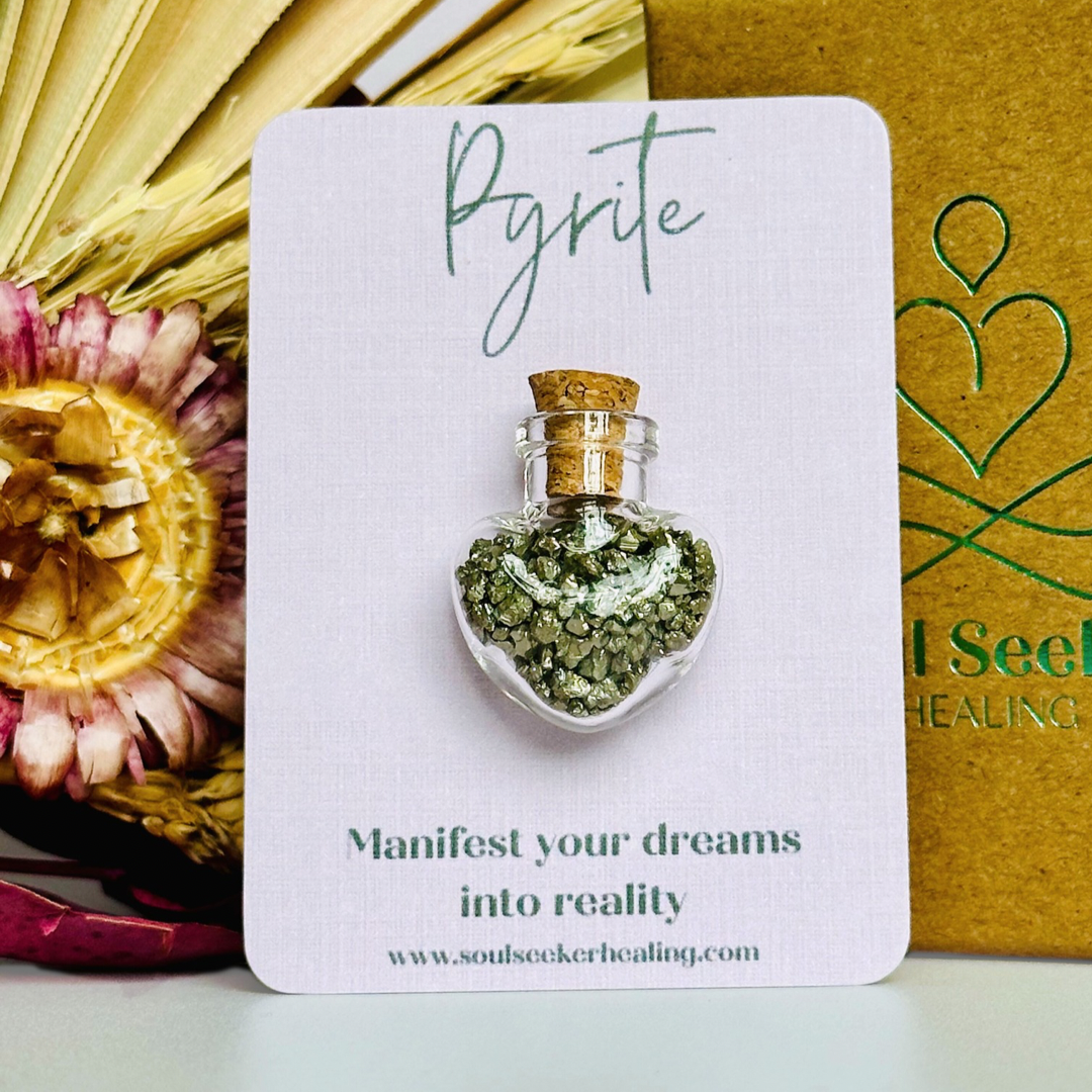 A pretty heart shaped mini glass bottle filled with powerful, ethically mined pyrite crystal chips, presented on an eco-friendly textured card with the words 'Manifest your dreams into reality.’
