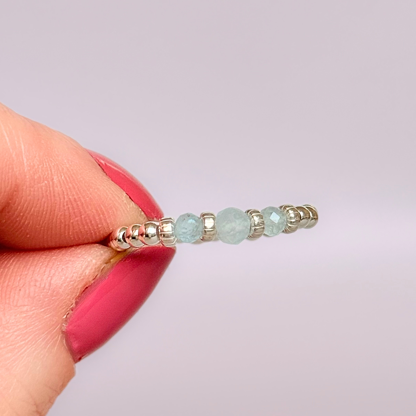 Aquamarine crystal trilogy sterling silver elasticated ring featuring three March birthstone gemstone faceted beads, on a band of sterling silver round beads. The crystals are seperated by sterling silver corrugated rondelles. The main crystal is 4mm and the ones at either side are 3mm. Pictured being held against a pink background.
