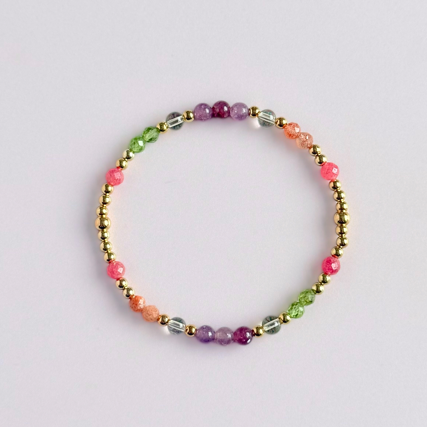 Dainty crystal bracelet featuring a carefully curated selection of genuine crystal beads inspired by wildflowers and free-spirited growth. Featuring 4mm smooth and faceted round gemstone beads with 3mm round accent beads in either sterling silver or gold vermeil. With peridot, sunstone, rhodochrosite, citrine and lepidolite. Pictured in gold, from above, against a light pink background.