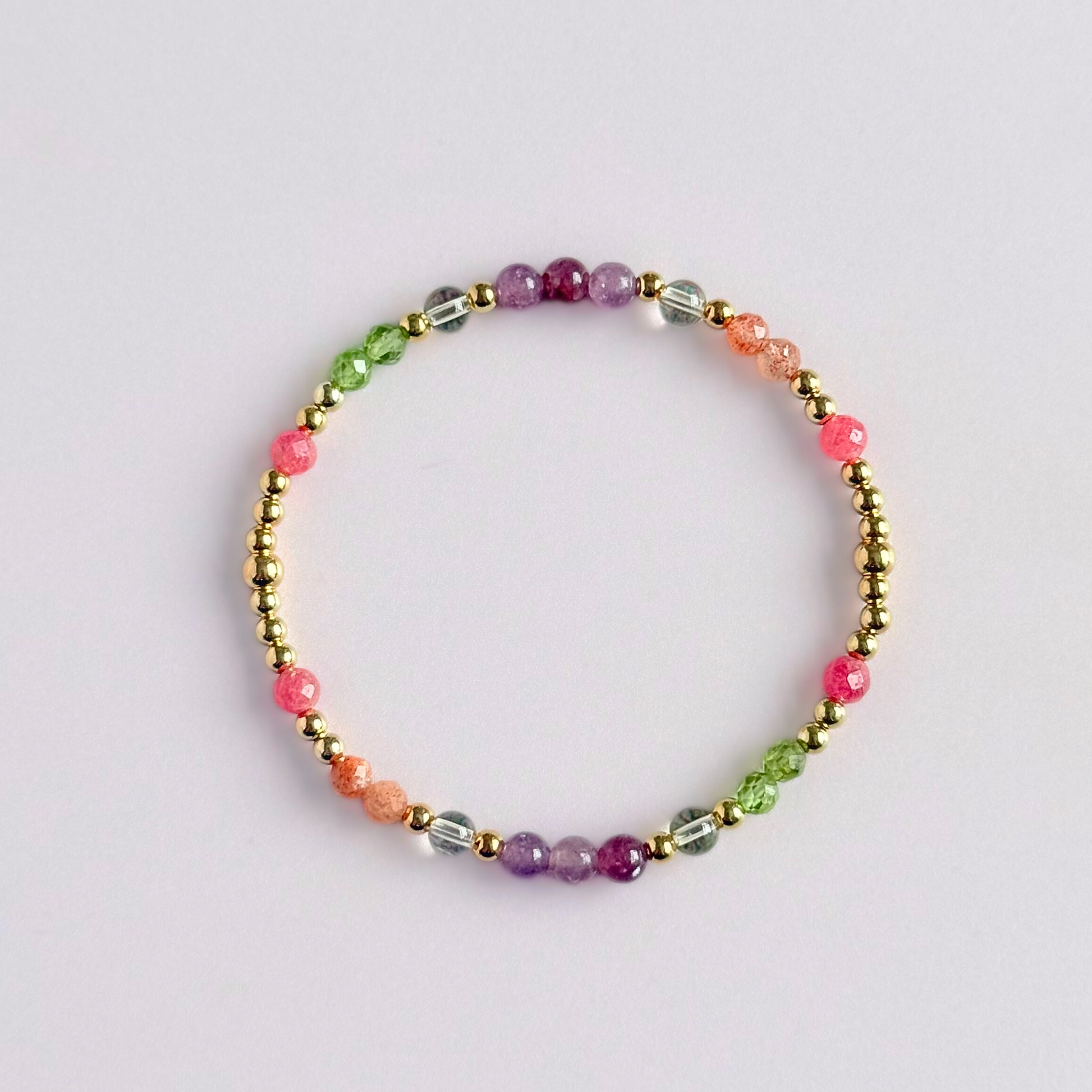 Dainty crystal bracelet featuring a carefully curated selection of genuine crystal beads inspired by wildflowers and free-spirited growth. Featuring 4mm smooth and faceted round gemstone beads with 3mm round accent beads in either sterling silver or gold vermeil. With peridot, sunstone, rhodochrosite, citrine and lepidolite. Pictured in gold, from above, against a light pink background.