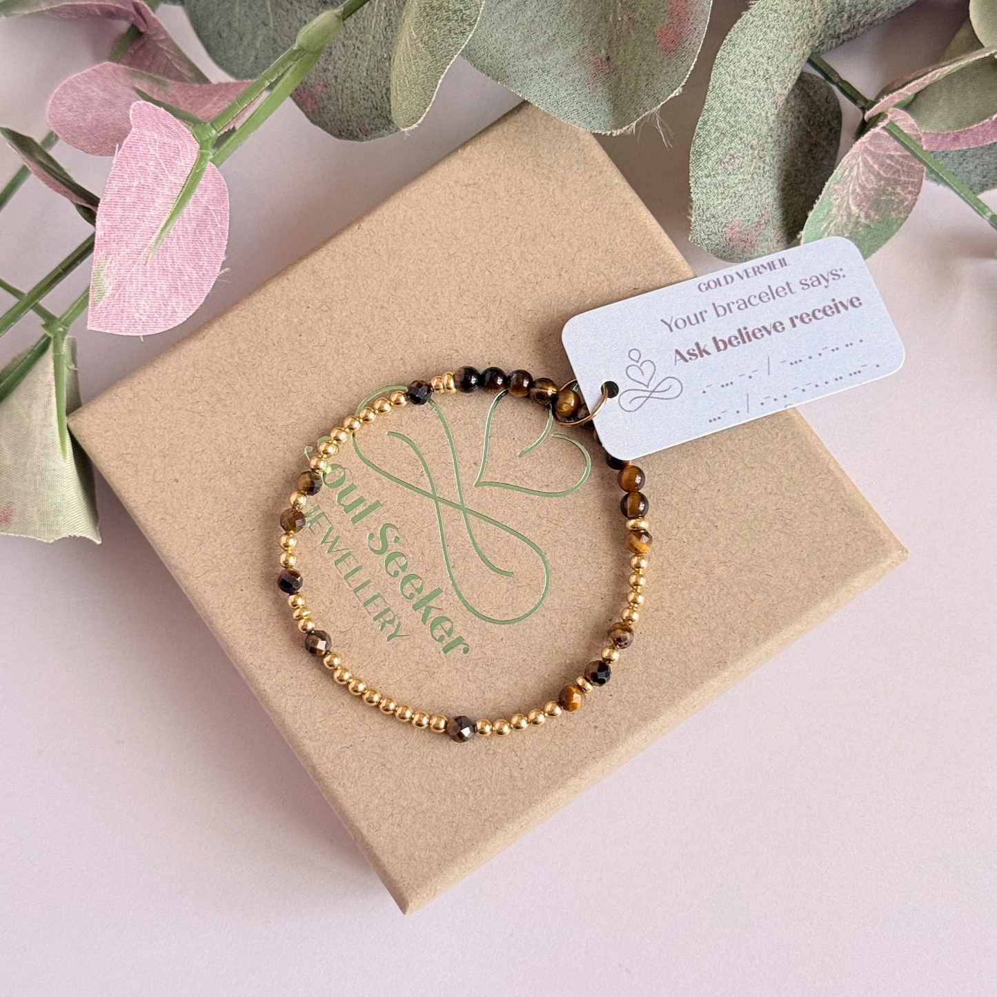 Tigers eye morse code bracelet that spells out the words ‘Ask, believe, receive’ in a combination of 4mm round smooth and faceted crystal beads with your choice of sterling silver or gold vermeil accent beads. Pictured in gold, on top of the gift box