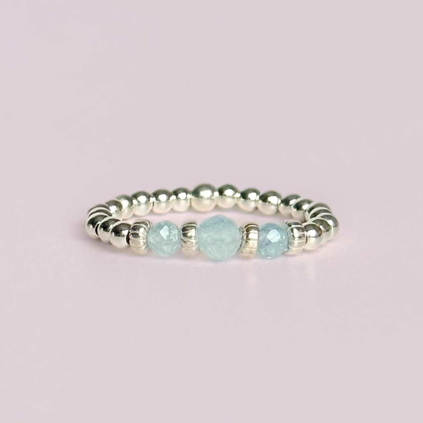 Aquamarine crystal trilogy sterling silver elasticated ring featuring three March birthstone gemstone faceted beads, on a band of sterling silver round beads. The crystals are seperated by sterling silver corrugated rondelles. The main crystal is 4mm and the ones at either side are 3mm. Shown flat, against a pink background.
