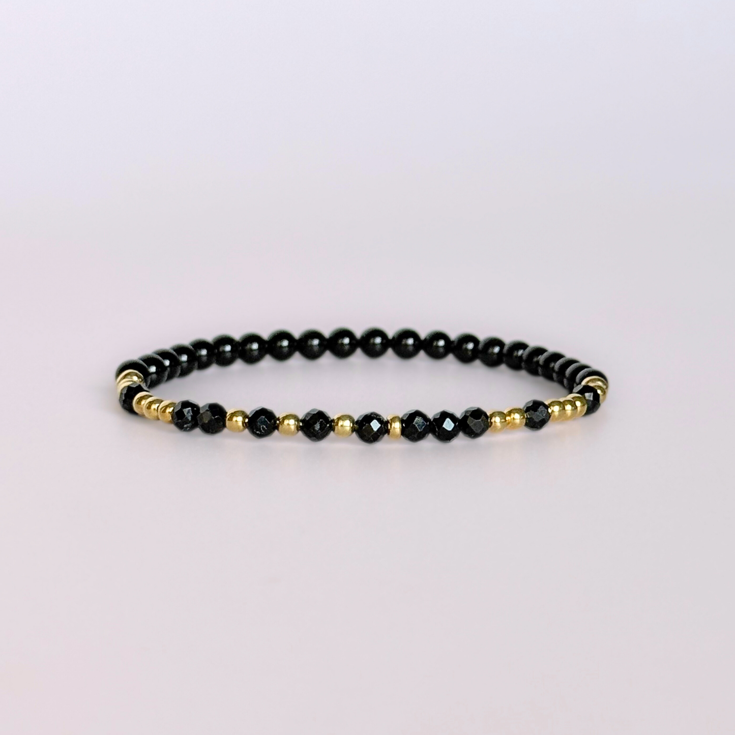 Elasticated black tourmaline crystal morse code bracelet spells out the words ‘Fuck off’ in a combination of 4mm round black tourmaline beads, 4mm faceted black tourmaline beads and either sterling silver or gold vermeil 3mm round beads. The words start and end at the rondelle spacer beads. Picture shows gold version against a light pink background.