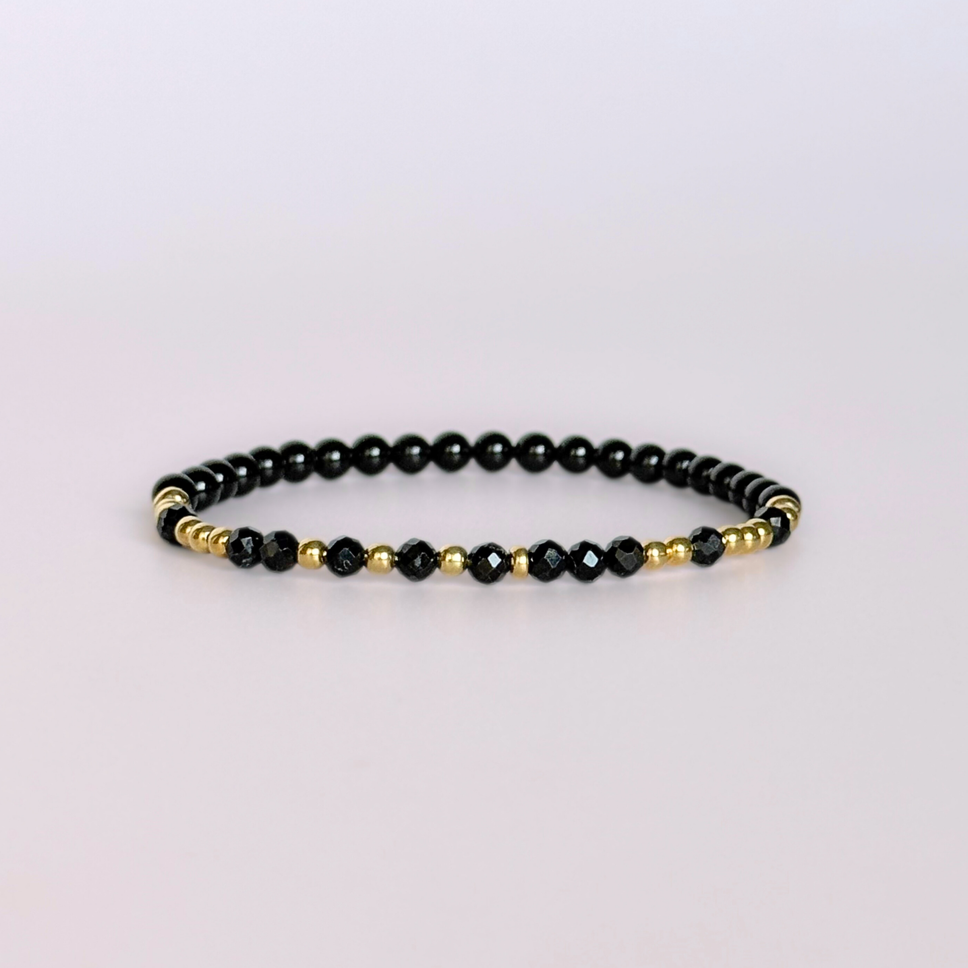 Elasticated black tourmaline crystal morse code bracelet spells out the words ‘Fuck off’ in a combination of 4mm round black tourmaline beads, 4mm faceted black tourmaline beads and either sterling silver or gold vermeil 3mm round beads. The words start and end at the rondelle spacer beads. Picture shows gold version against a light pink background.