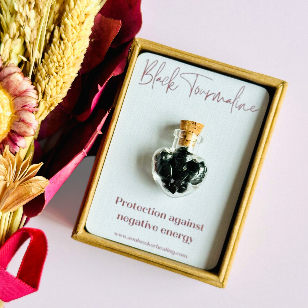 A pretty heart shaped mini glass bottle filled with powerful, ethically mined black tourmaline crystal chips, presented on an eco-friendly textured card with the words 'Protection against negative energy.’