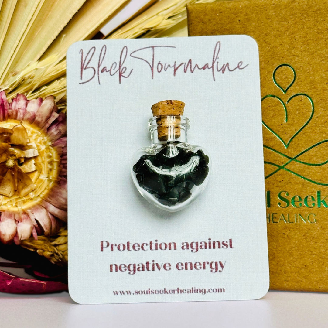 A pretty heart shaped mini glass bottle filled with powerful, ethically mined black tourmaline crystal chips, presented on an eco-friendly textured card with the words 'Protection against negative energy.’