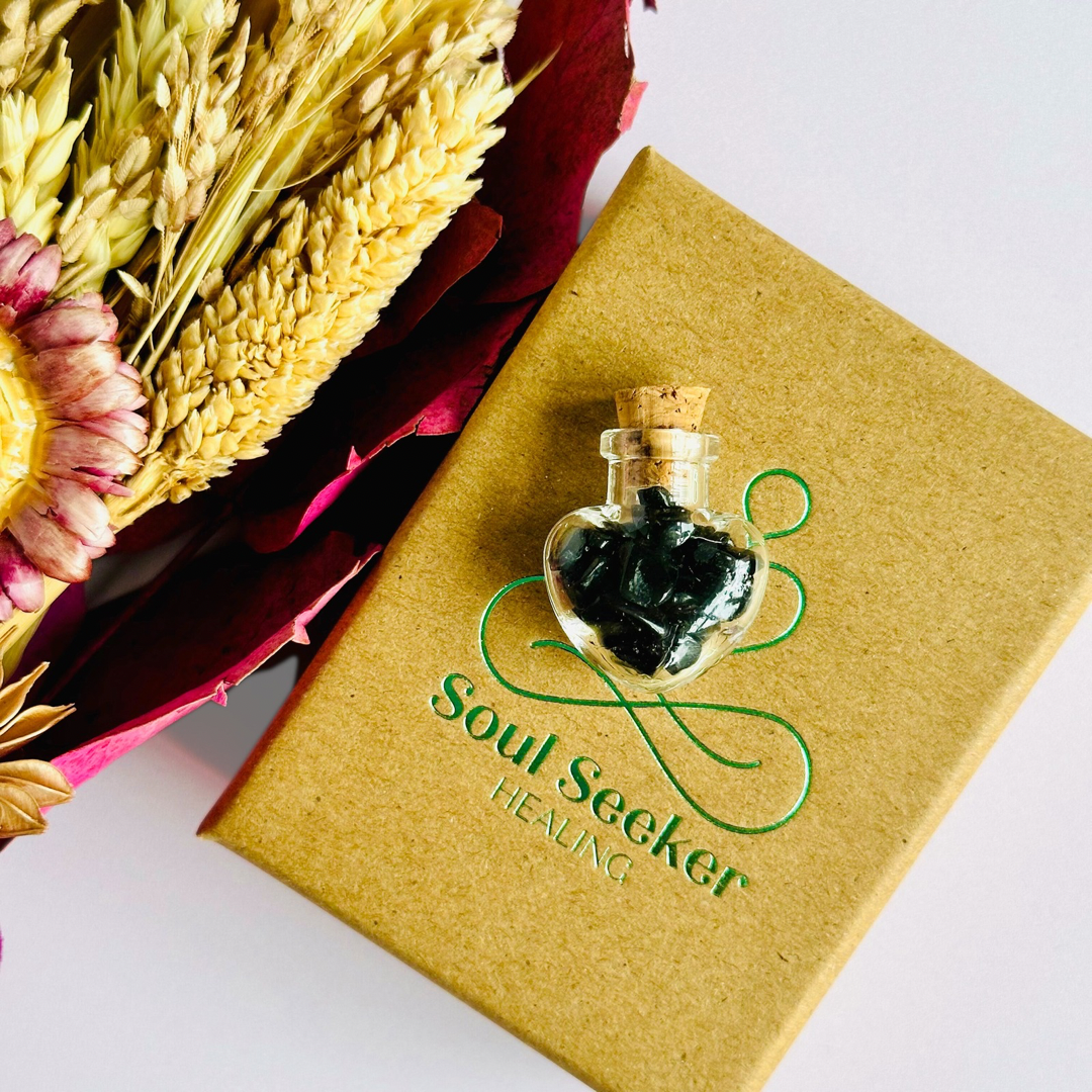 A pretty heart shaped mini glass bottle filled with powerful, ethically mined black tourmaline crystal chips, presented on an eco-friendly textured card with the words 'Protection against negative energy.’