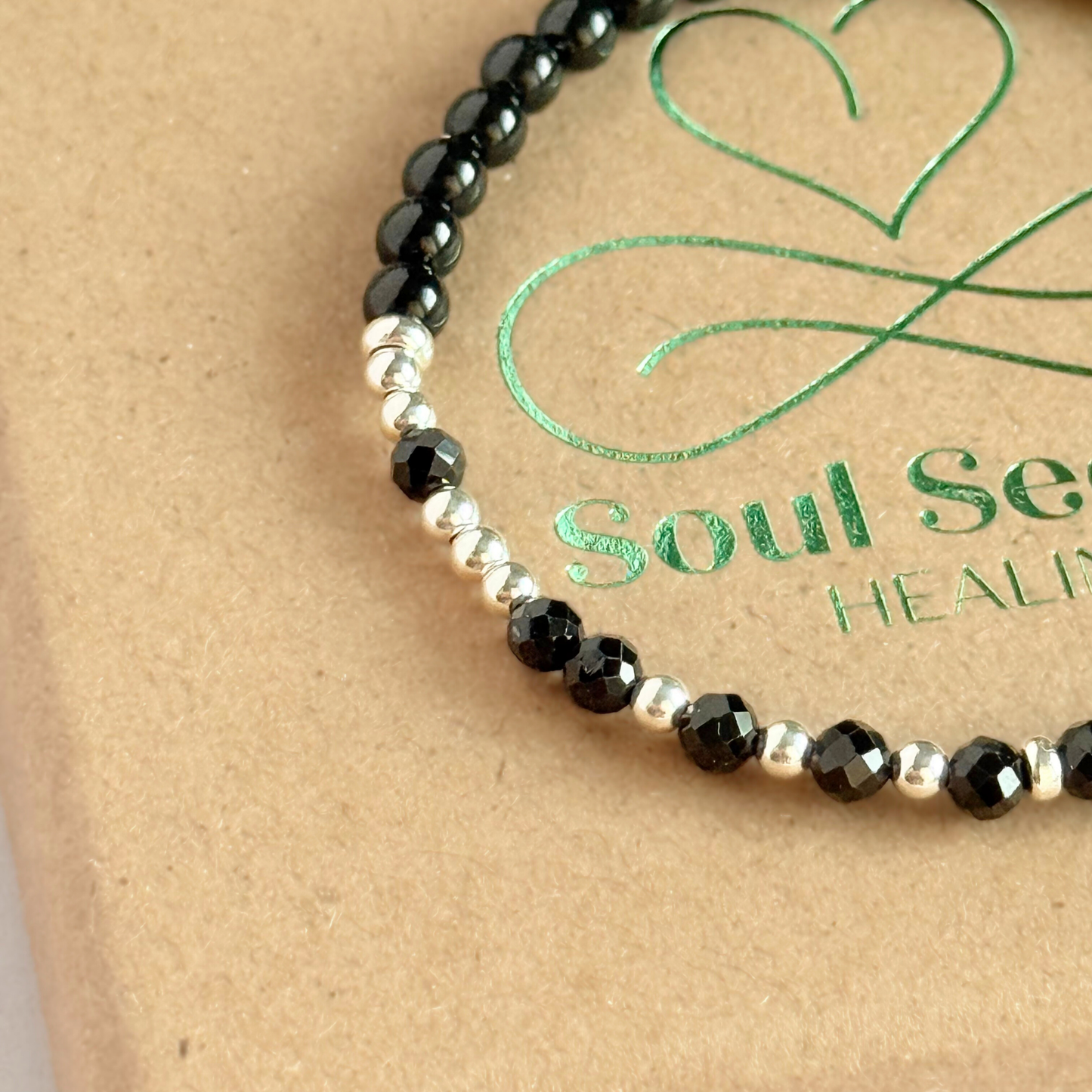 Elasticated black tourmaline crystal morse code bracelet spells out the words ‘Fuck off’ in a combination of 4mm round black tourmaline beads, 4mm faceted black tourmaline beads and either sterling silver or gold vermeil 3mm round beads. The words start and end at the rondelle spacer beads. Picture shows a close up of the silver version.
