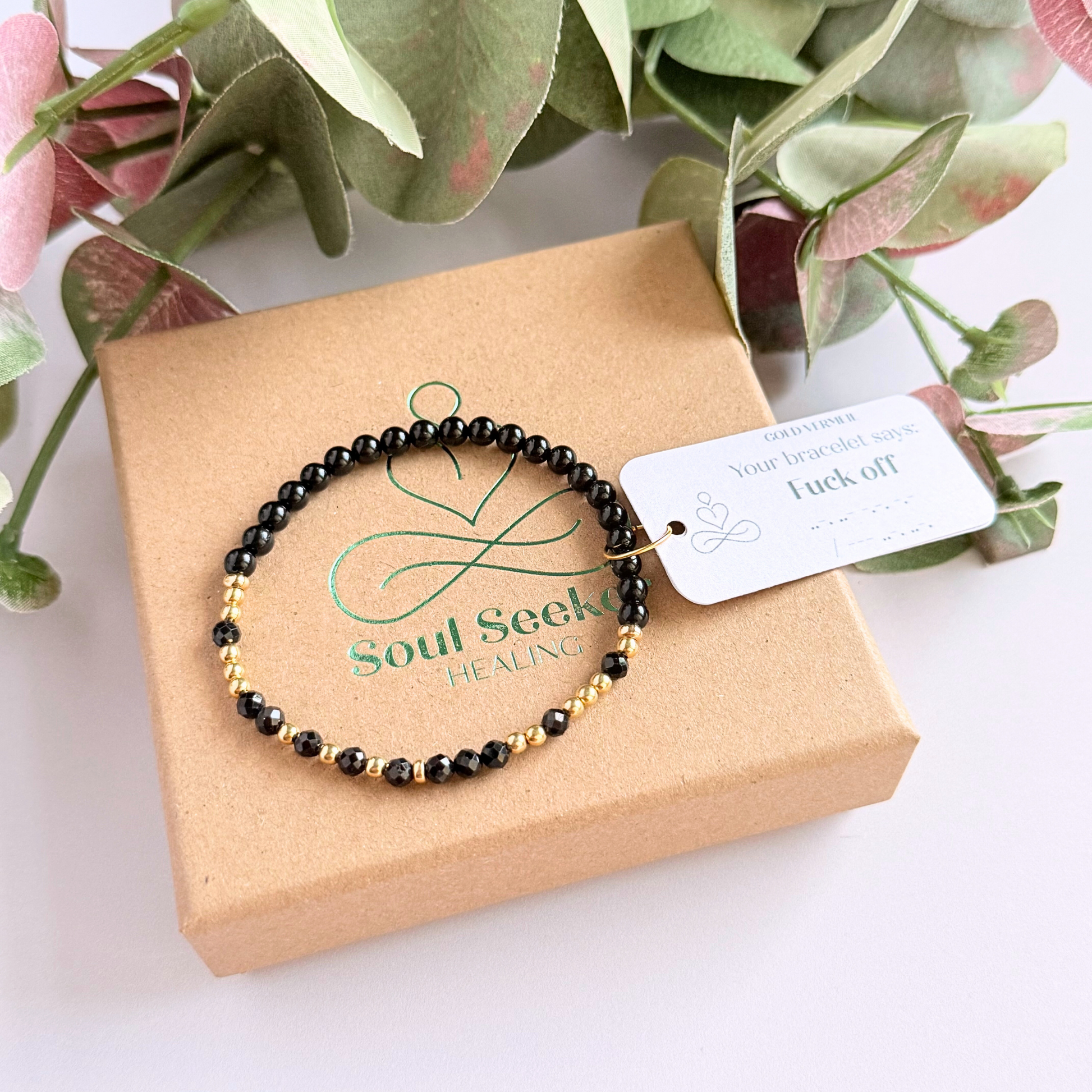 Elasticated black tourmaline crystal morse code bracelet spells out the words ‘Fuck off’ in a combination of 4mm round black tourmaline beads, 4mm faceted black tourmaline beads and either sterling silver or gold vermeil 3mm round beads. The words start and end at the rondelle spacer beads. Picture shows gold version on top of the gift box.